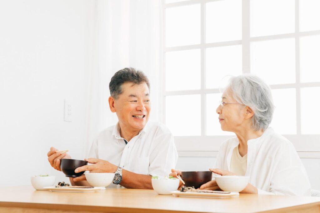 明るいダイニングで、笑顔で会話を楽しみながら和食を食べる高齢の夫婦の様子