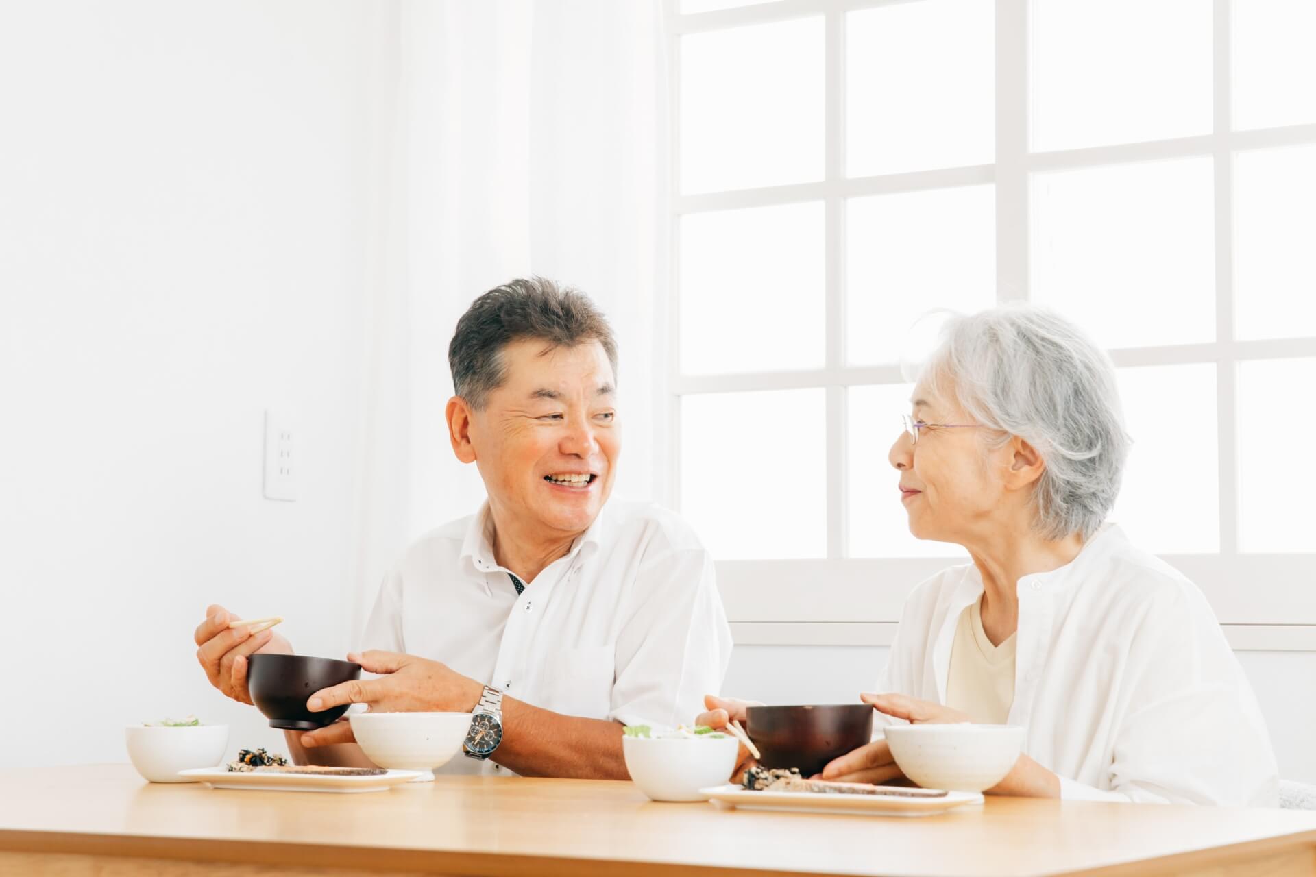 明るいダイニングで、笑顔で会話を楽しみながら和食を食べる高齢の夫婦の様子