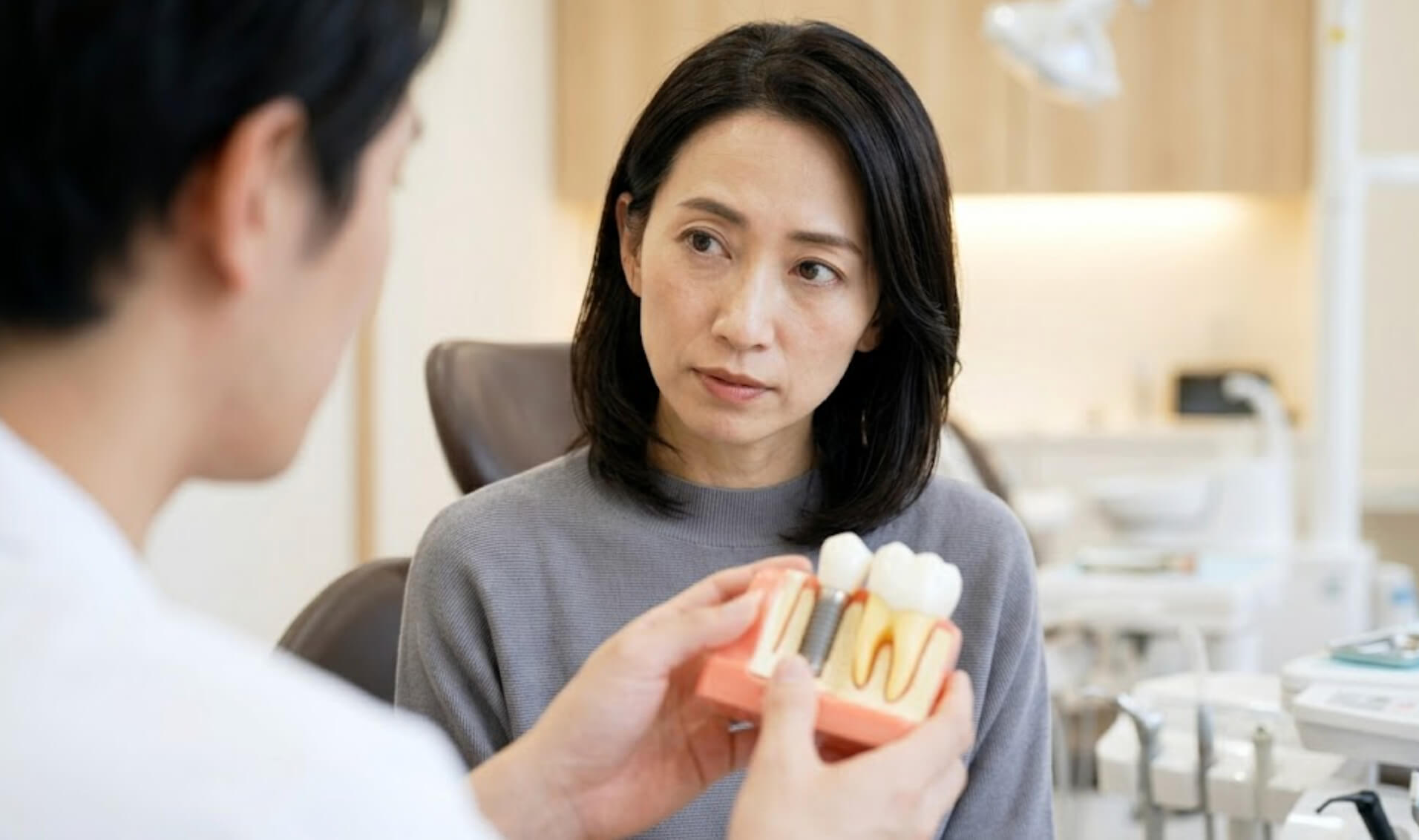 歯科医院で歯科医師からインプラントの模型を使って説明を受ける、真剣な表情の日本人女性の様子