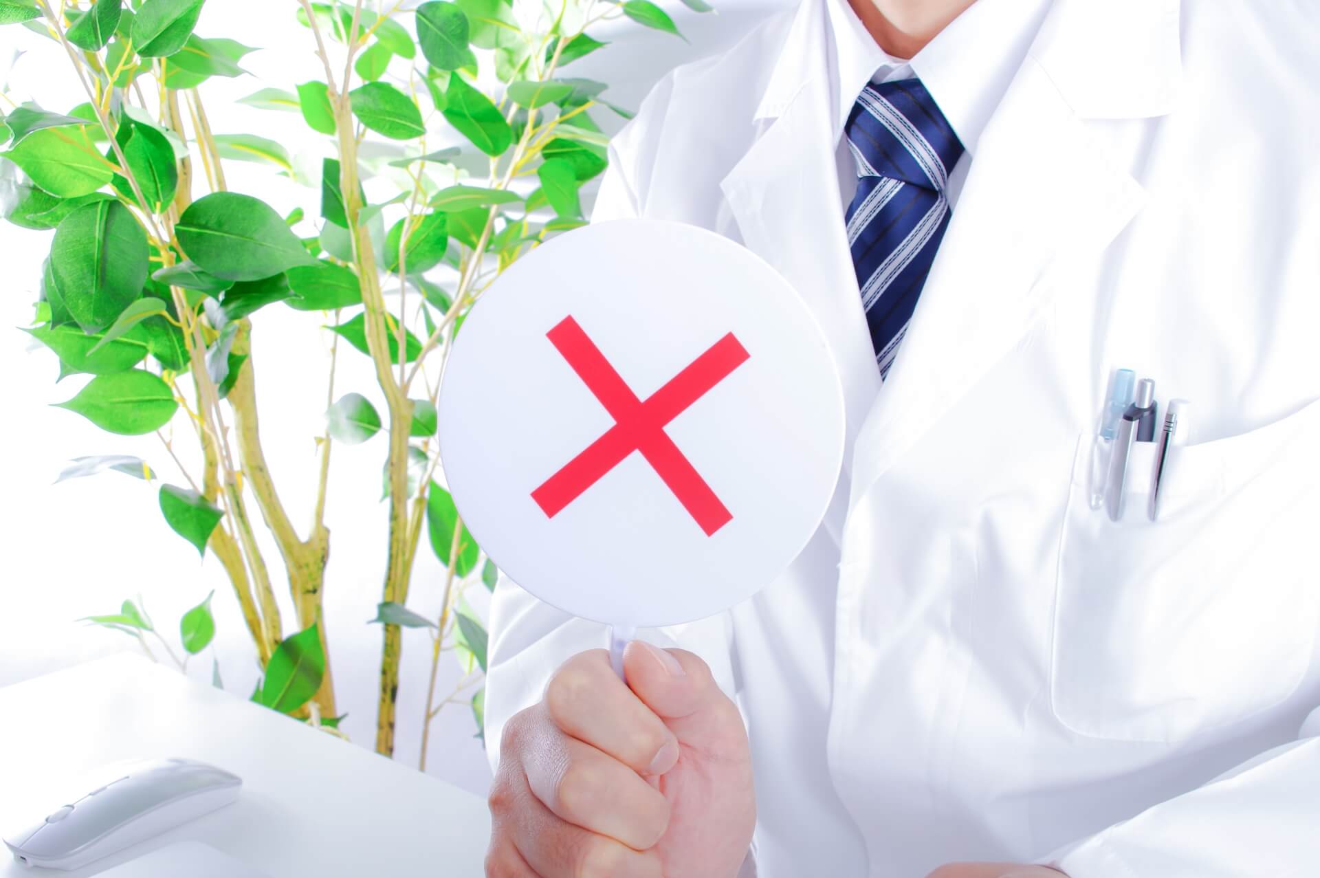 白衣姿の歯科医師が保険適用外であることを表すバツ印のプレートを掲げる様子